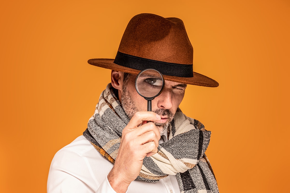 Male detective with magnifying glass, wearing fashionable scarf and brown hat, posing on yellow studio background. Portrait of handsome man with beard.