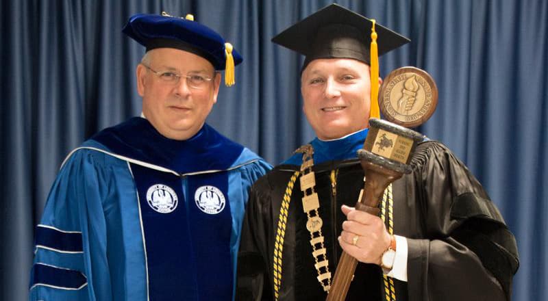 Dr. Burckel of McNeese stands with Dr. Henderson of University of Louisiana System wearing graduation regalia and holding the made