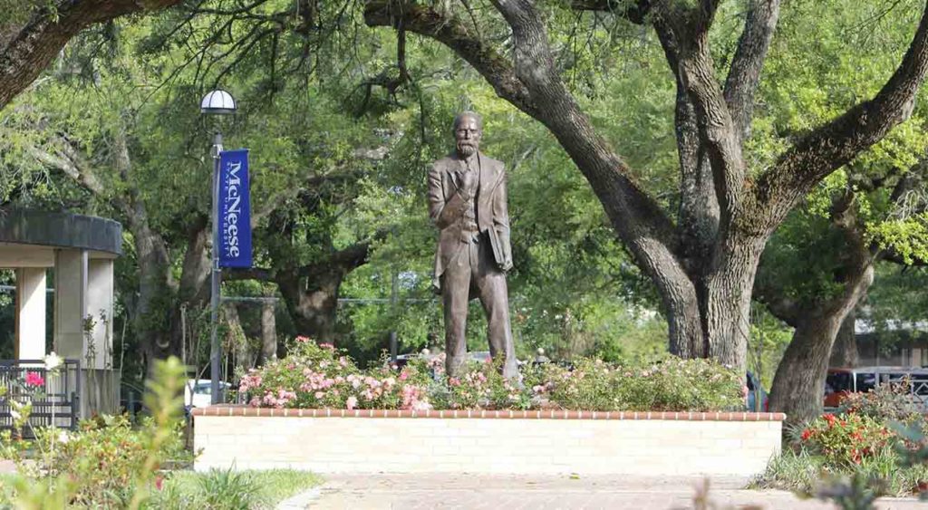 A statue of John McNeese surrounded by greenery and flowers on campus