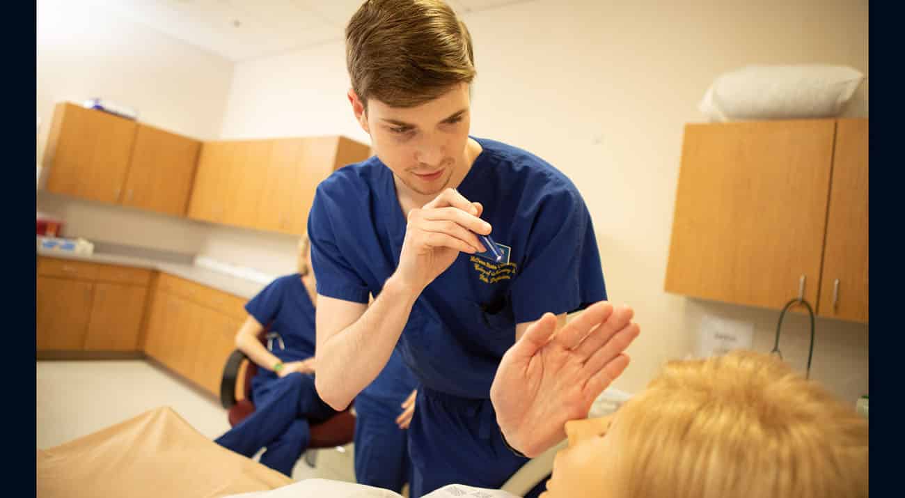 A male nursing student checks the eye reflex of a simulated patient.