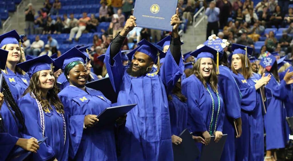 A McNeese graduate holds his diploma cover high as his peers look on.