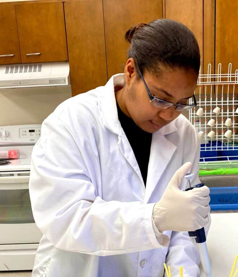 Scientist wearing a lab coat and gloves using a pipette in a laboratory setting.