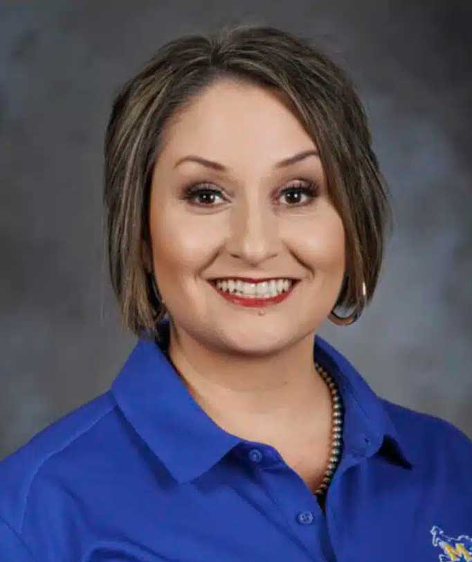 Smiling woman with short hair wearing a blue polo shirt against a gray background.