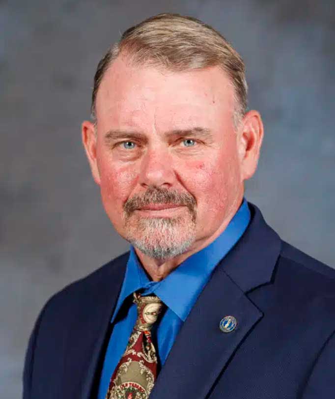 Man in formal blue suit with paisley tie, grey beard and short hair, facing camera in professional portrait setting.
