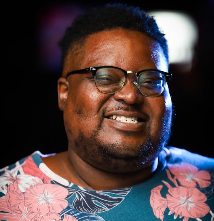 Smiling man with glasses wearing a floral shirt against a dark background