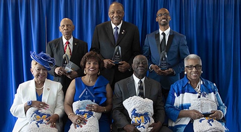 Seven people in formal attire holding awards and blankets with blue backdrop.