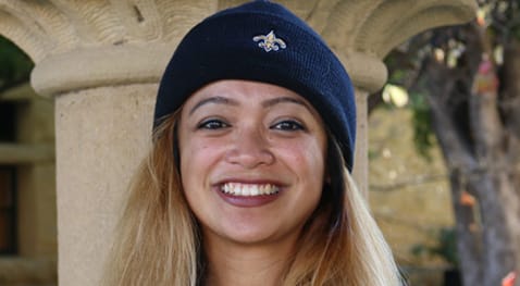 Person smiling outdoors wearing a beanie with a small embroidered emblem, standing in front of a stone column.