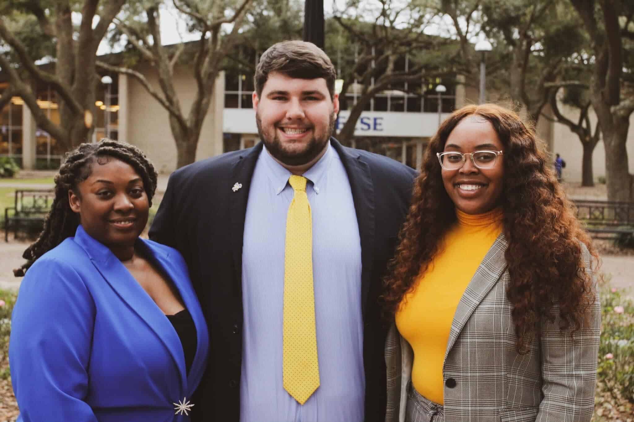 Three smiling people in professional attire outdoors in front of a building with trees.
