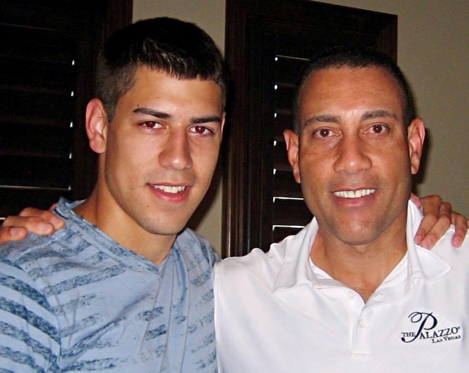 Two smiling men standing together indoors, one in a blue striped shirt and the other in a white Palazzo polo.