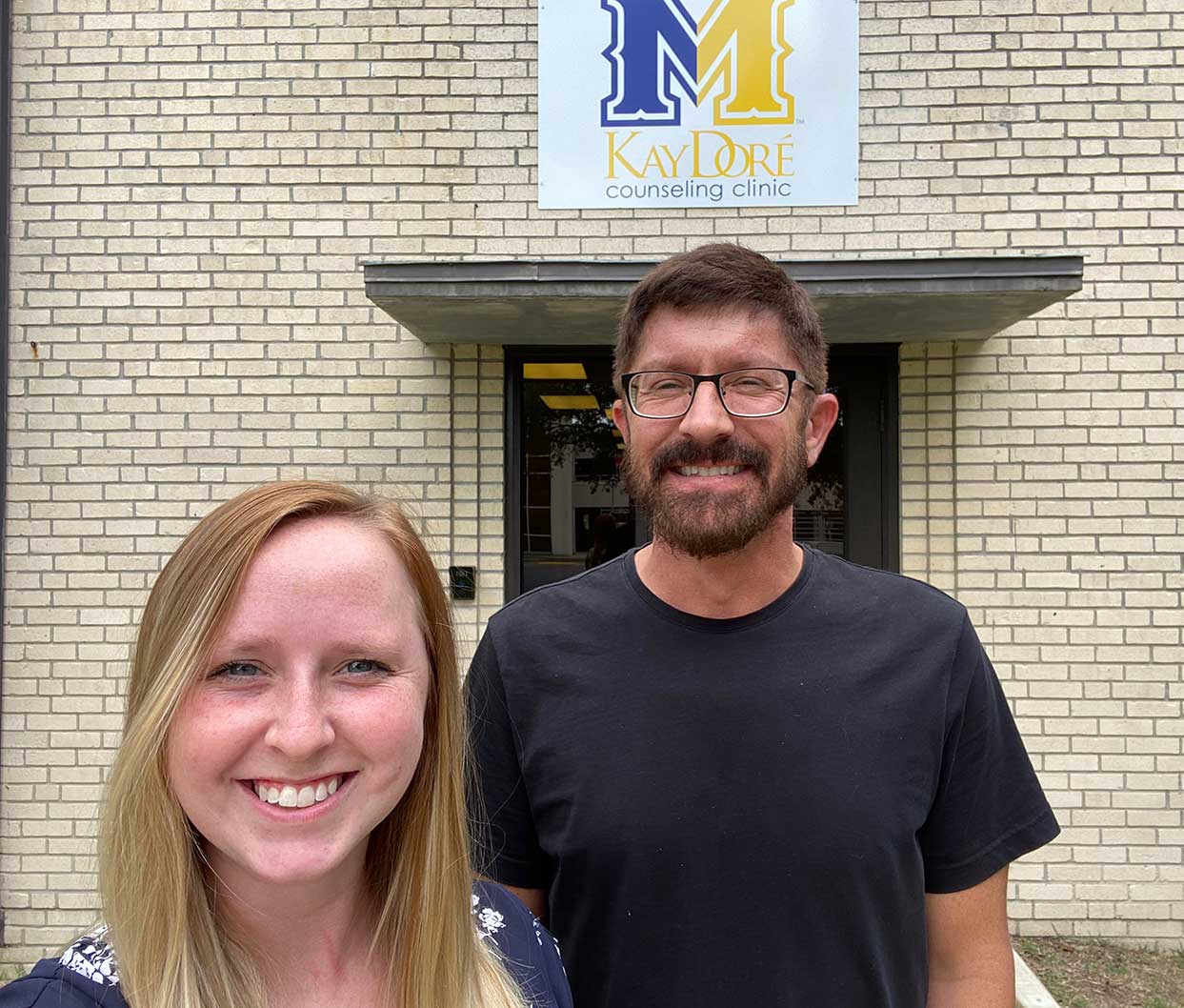 Two people smiling outside Kay Doré Counseling Clinic building with sign visible.