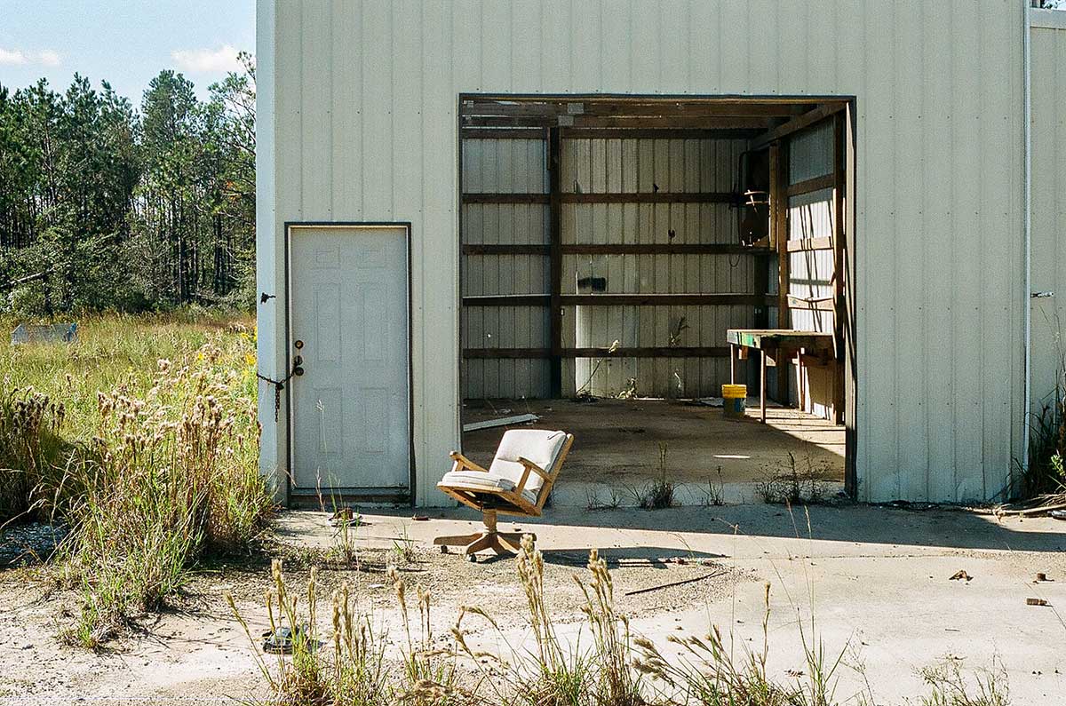 Abandoned warehouse with open door, empty chair outside, overgrown grass, and forest background