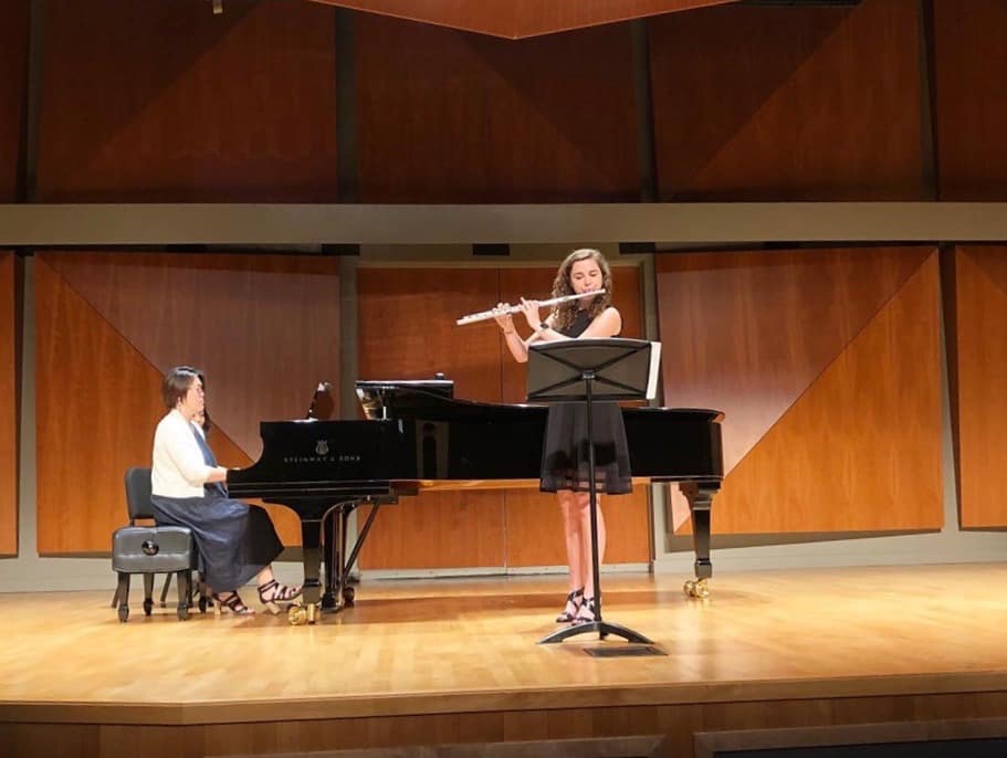 Flutist performs with pianist on stage in concert hall with wooden backdrop.