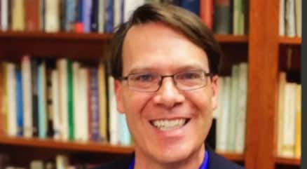 Smiling person wearing glasses in a library with shelves of books in the background.
