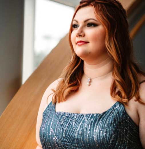 Woman in a blue shimmer gown with red hair, looking out a window, in a softly lit room.