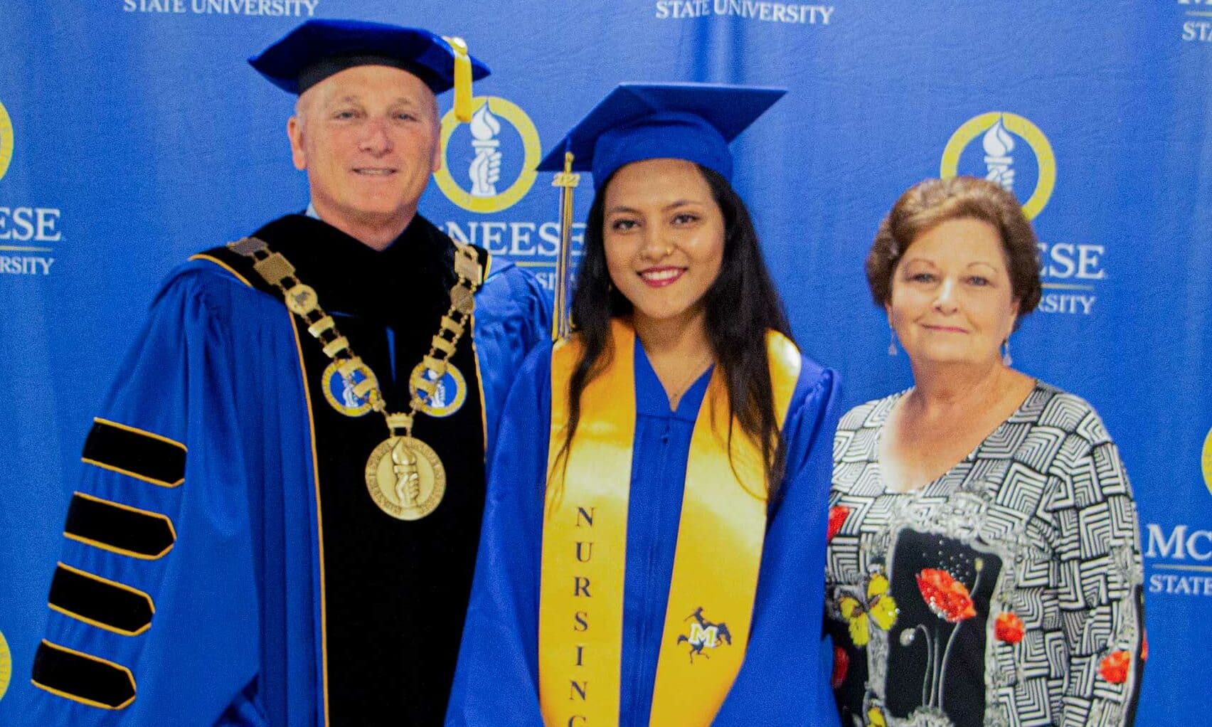 Graduate in blue cap and gown with honors sashes, stands with university official and family member at ceremony.
