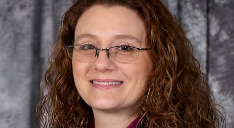 Smiling woman with curly brown hair and glasses in front of gray curtain background