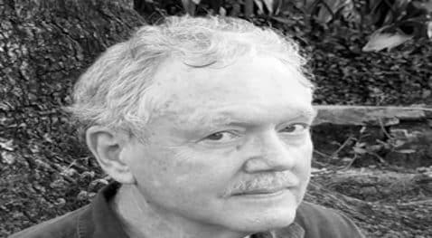 Elderly man with gray hair and mustache sitting outdoors near a tree