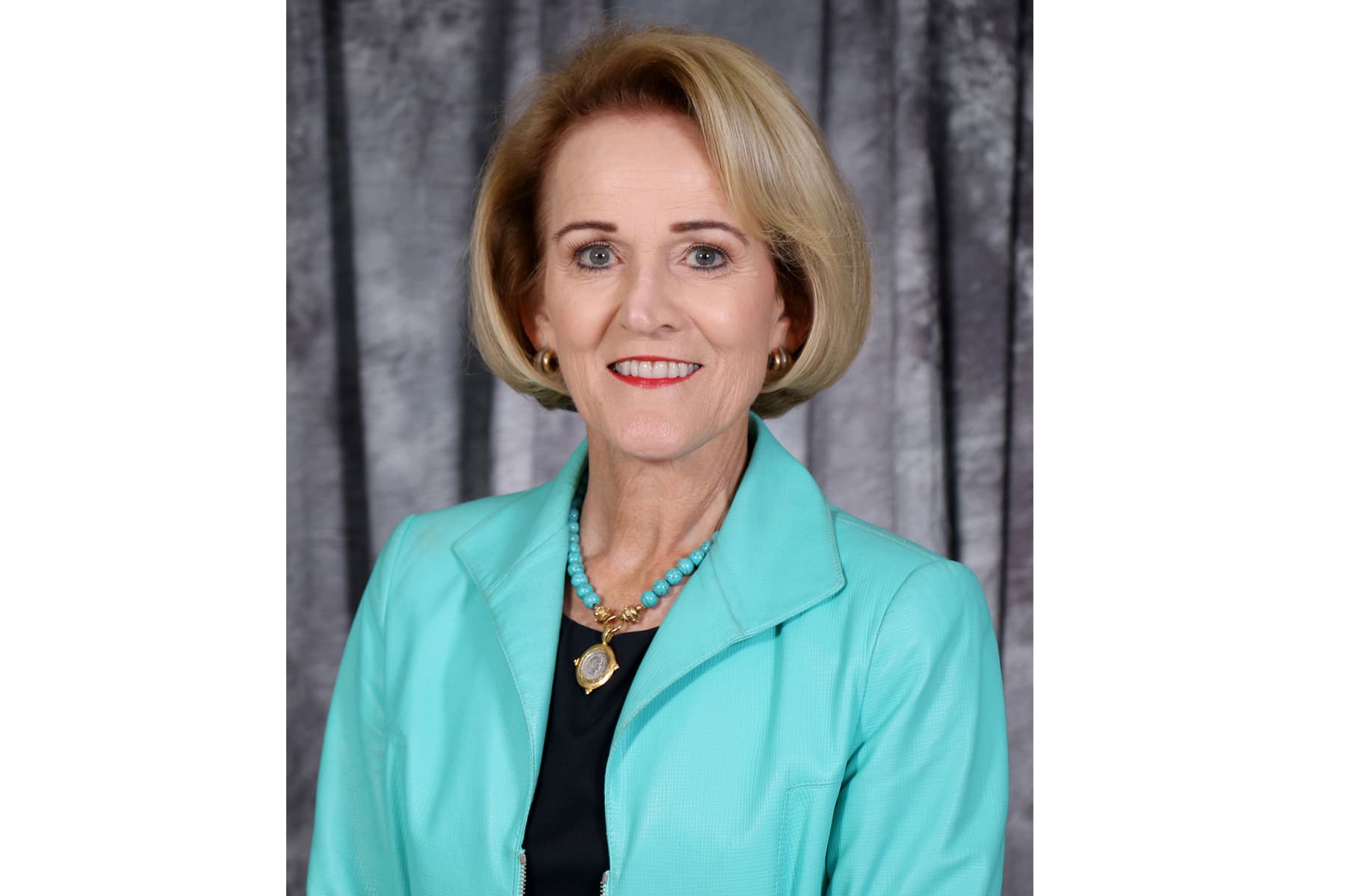Smiling woman with short blonde hair wearing turquoise blazer and matching necklace, gray backdrop.