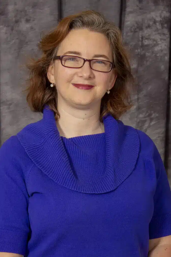 Smiling woman with glasses in blue sweater against a gray background.