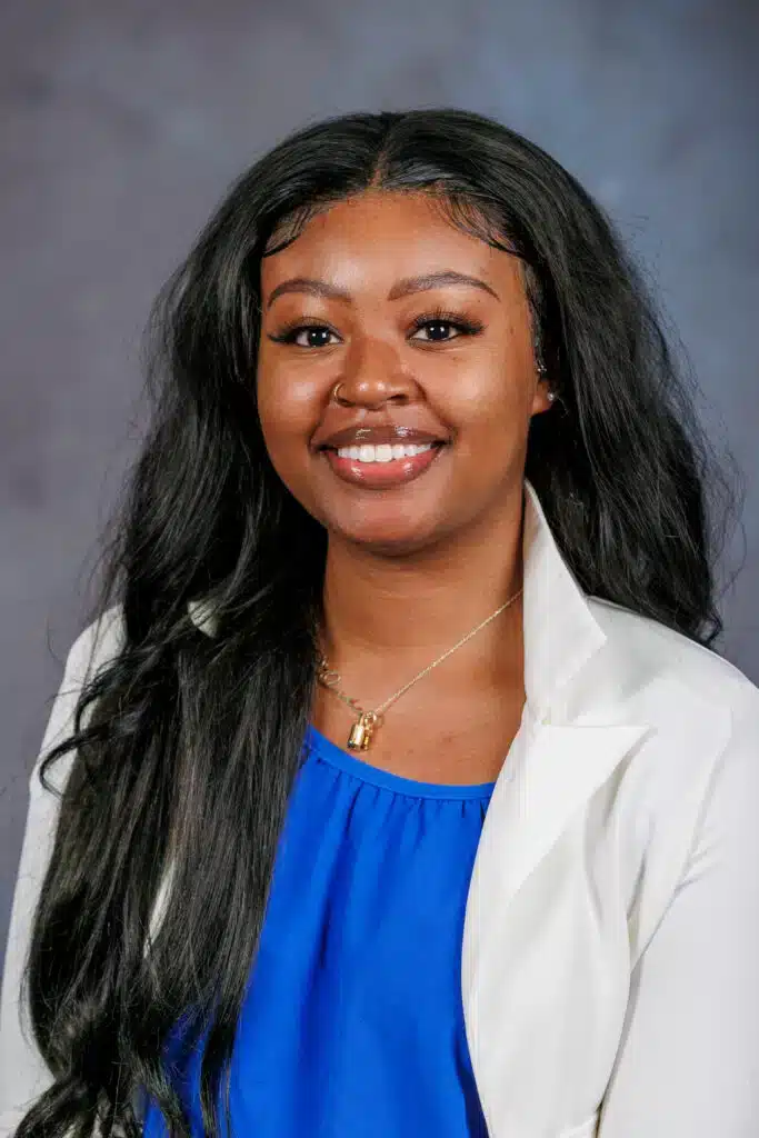 Smiling woman with long black hair in a white blazer and blue top against a gray background.