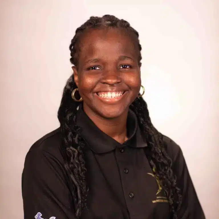 Smiling woman with braided hair wearing a black polo shirt with gold embroidery, neutral background.
