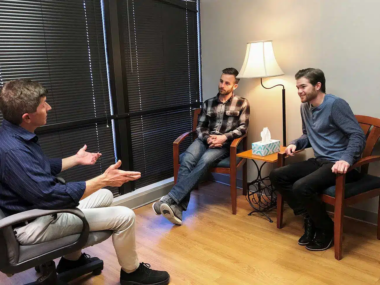 Three men having a discussion in a small office with a lamp and tissue box on a side table