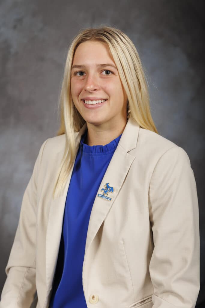 Smiling woman with long blonde hair in a beige blazer and blue shirt against a grey background.