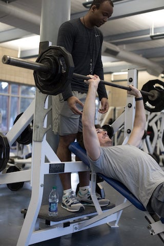 Man bench pressing weights with trainer assisting in gym setting.