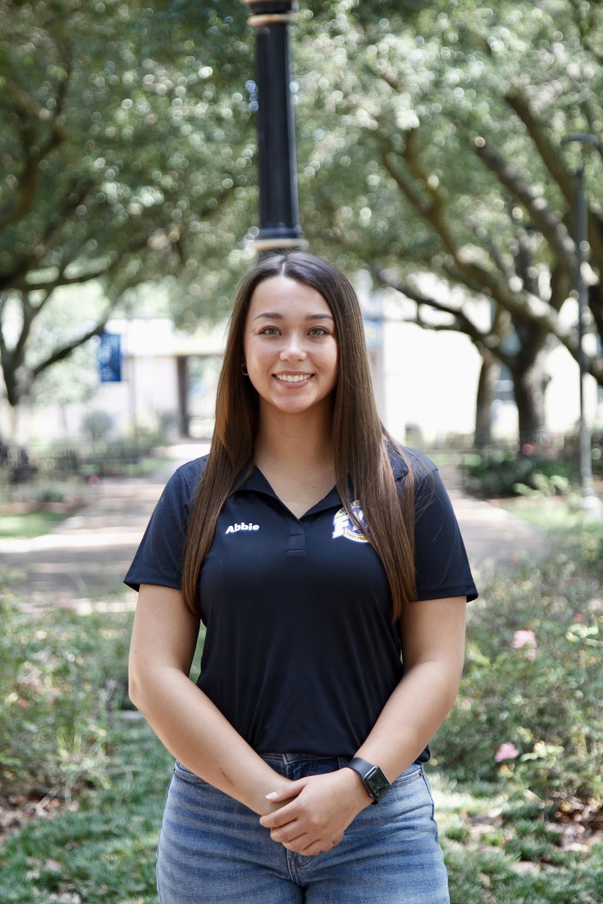 Woman smiling in a park setting, wearing a black polo shirt and jeans, surrounded by trees and greenery.