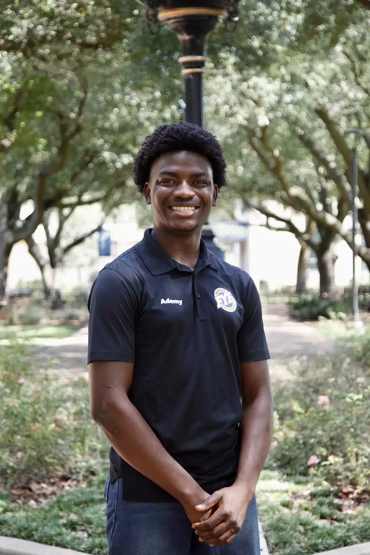 Smiling person standing in a garden wearing a black polo shirt with trees in the background.