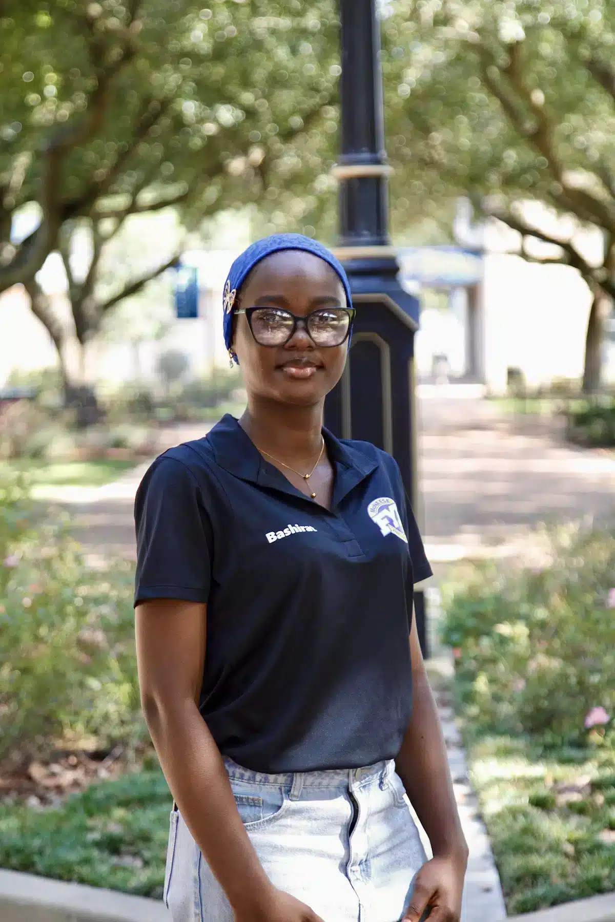 Smiling person with glasses and blue headscarf outdoors, wearing a black polo shirt and jeans.