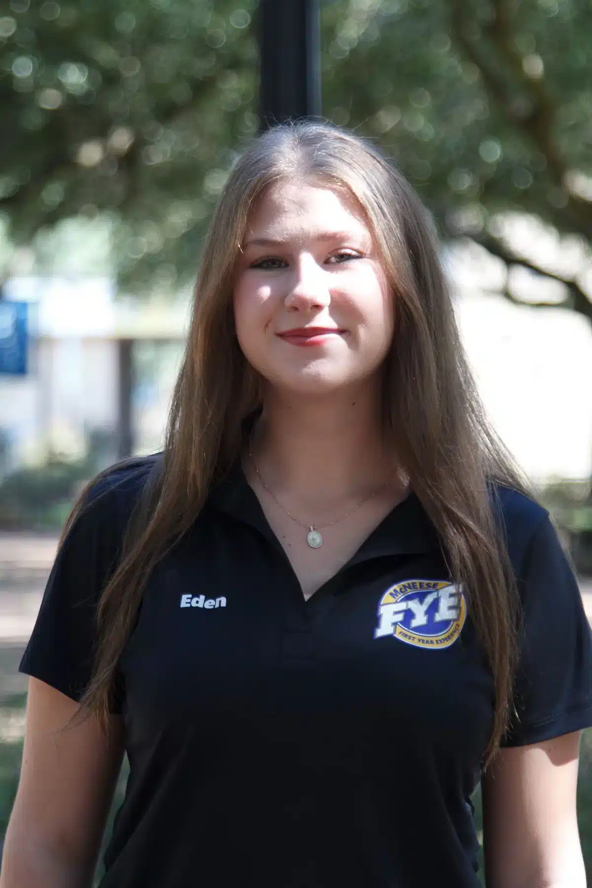 Woman with long hair wearing a black FYE McNeese polo shirt, outdoors with blurred leafy background.