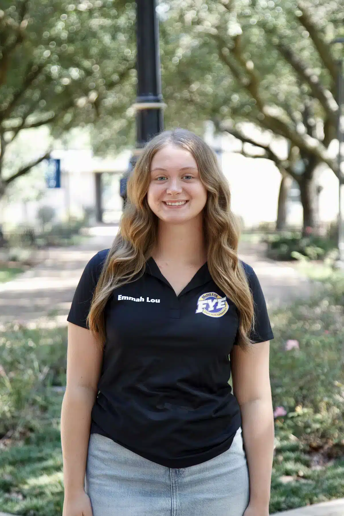 Young woman in black FYE polo smiling outdoors with trees in the background.