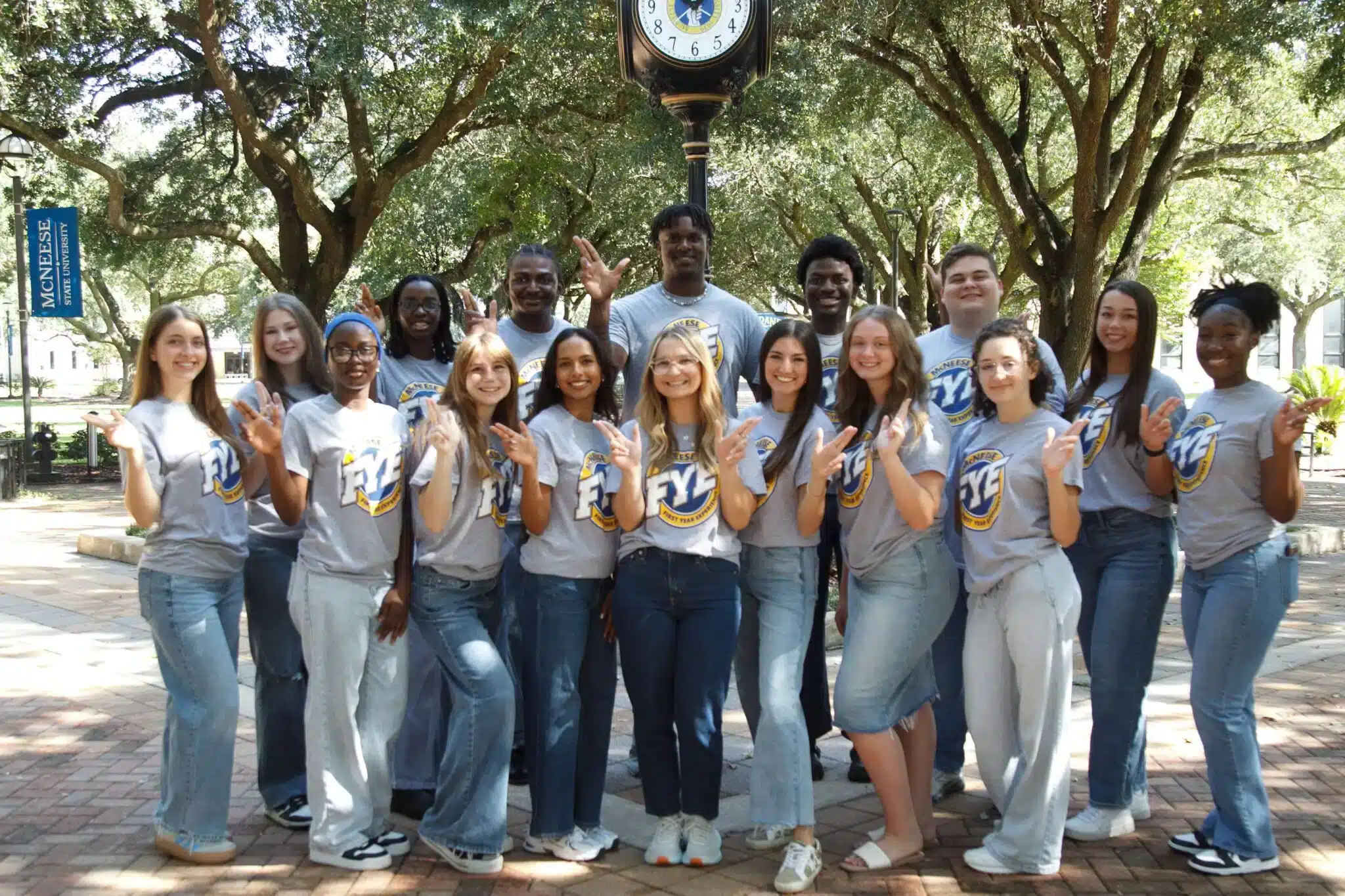 Group of students in McNeese State University shirts posing under a clock in a tree-lined campus walkway.