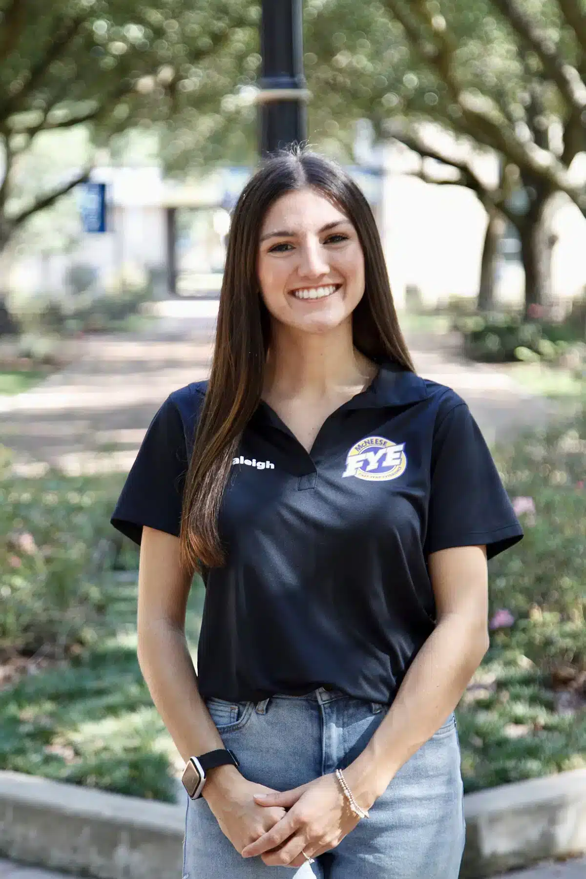 Smiling woman in black shirt with logo stands in a park, hands clasped, trees in the background.