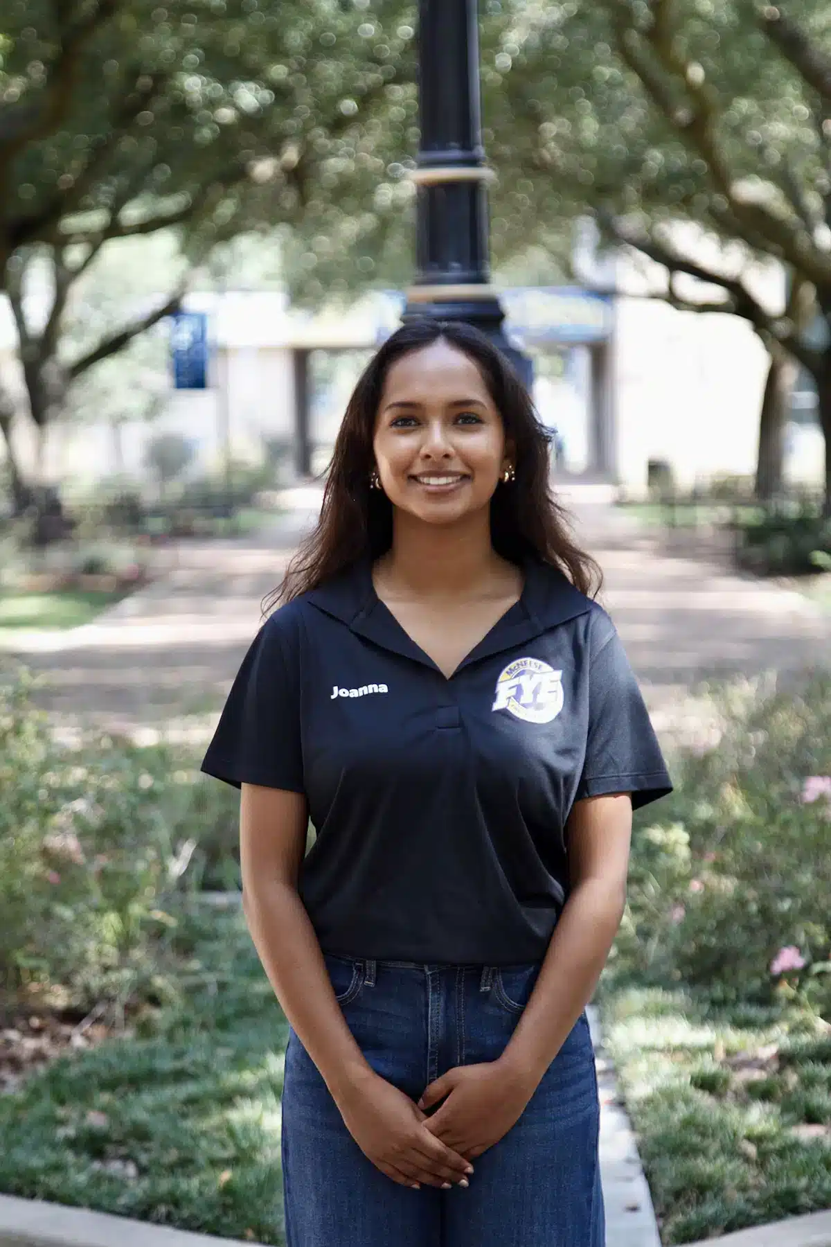 Smiling woman in black polo shirt standing outdoors on a sunny day, with trees in the background.