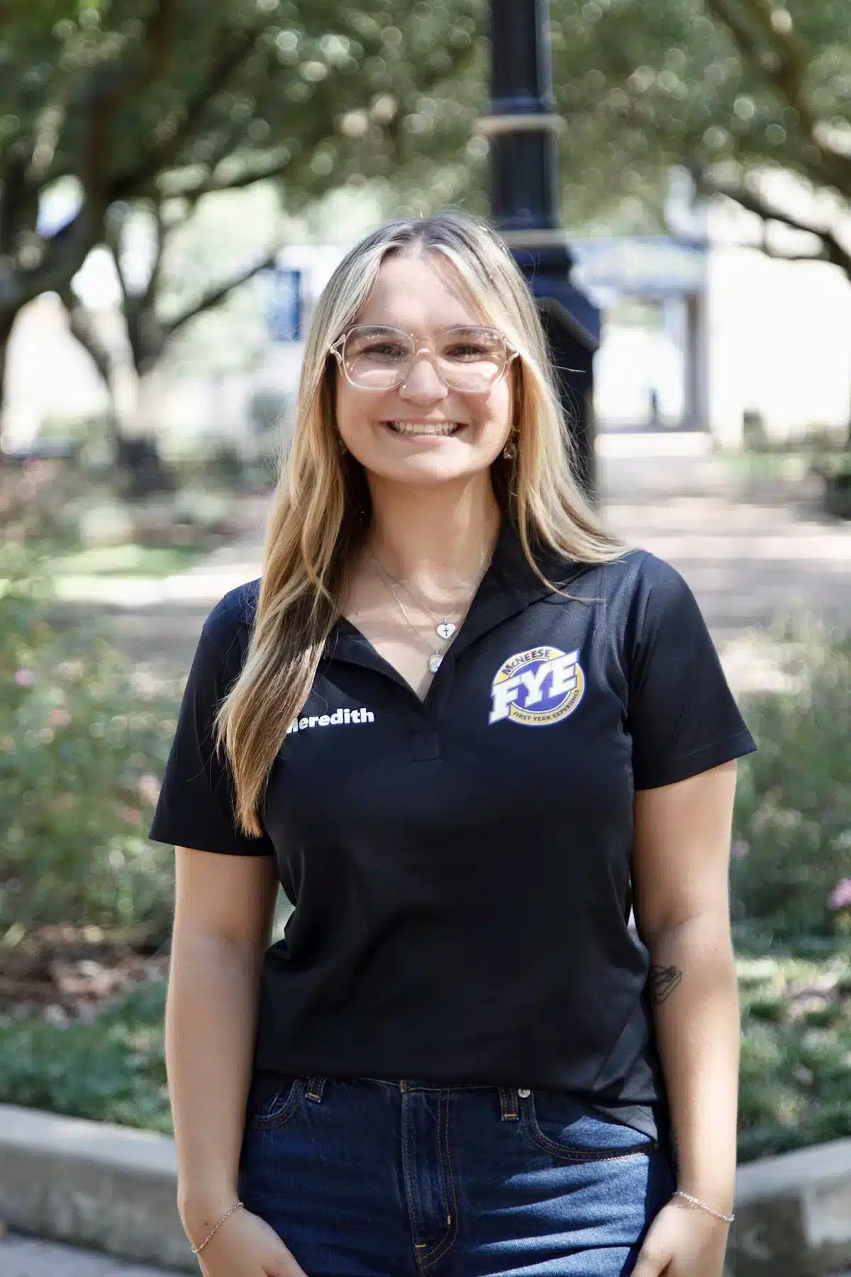 Smiling woman wearing a McNeese State University FYE polo, outdoors in a park with trees in the background.