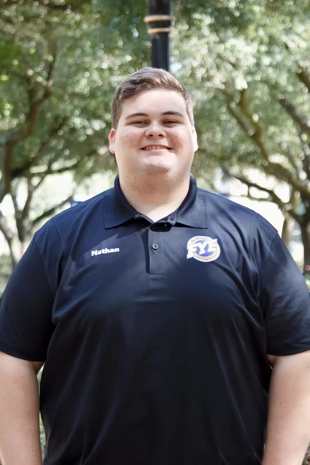 Smiling person in a navy FYE shirt outdoors with trees in the background