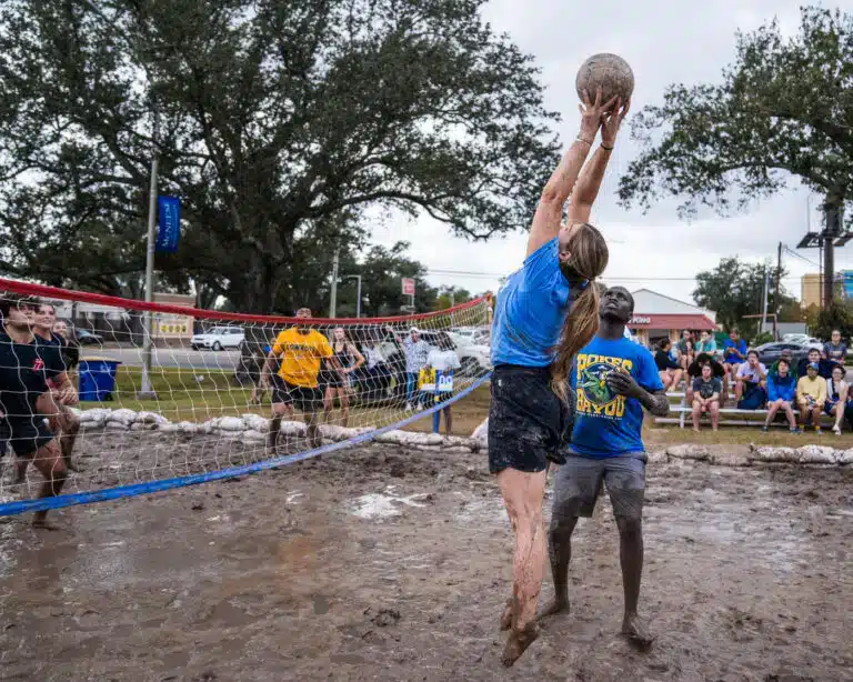 Players competing in a muddy outdoor volleyball game, jumping to spike the ball over a net.