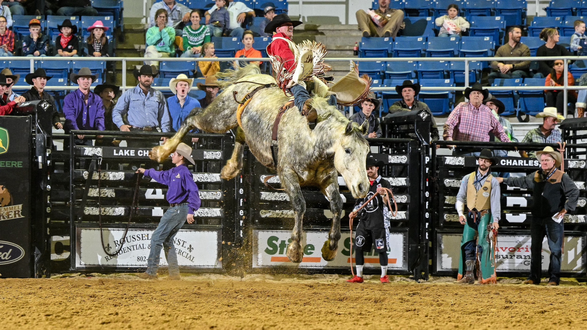 Rodeo cowboy riding bucking horse in arena surrounded by spectators and staff.