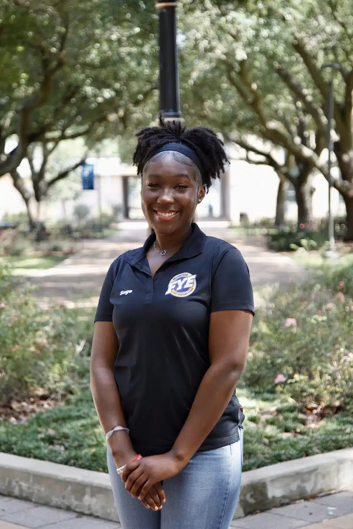 Smiling woman in black polo shirt with logo outdoors in a leafy park.