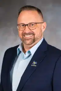 Smiling man in glasses wearing a blue suit with McNeese State University pin on lapel