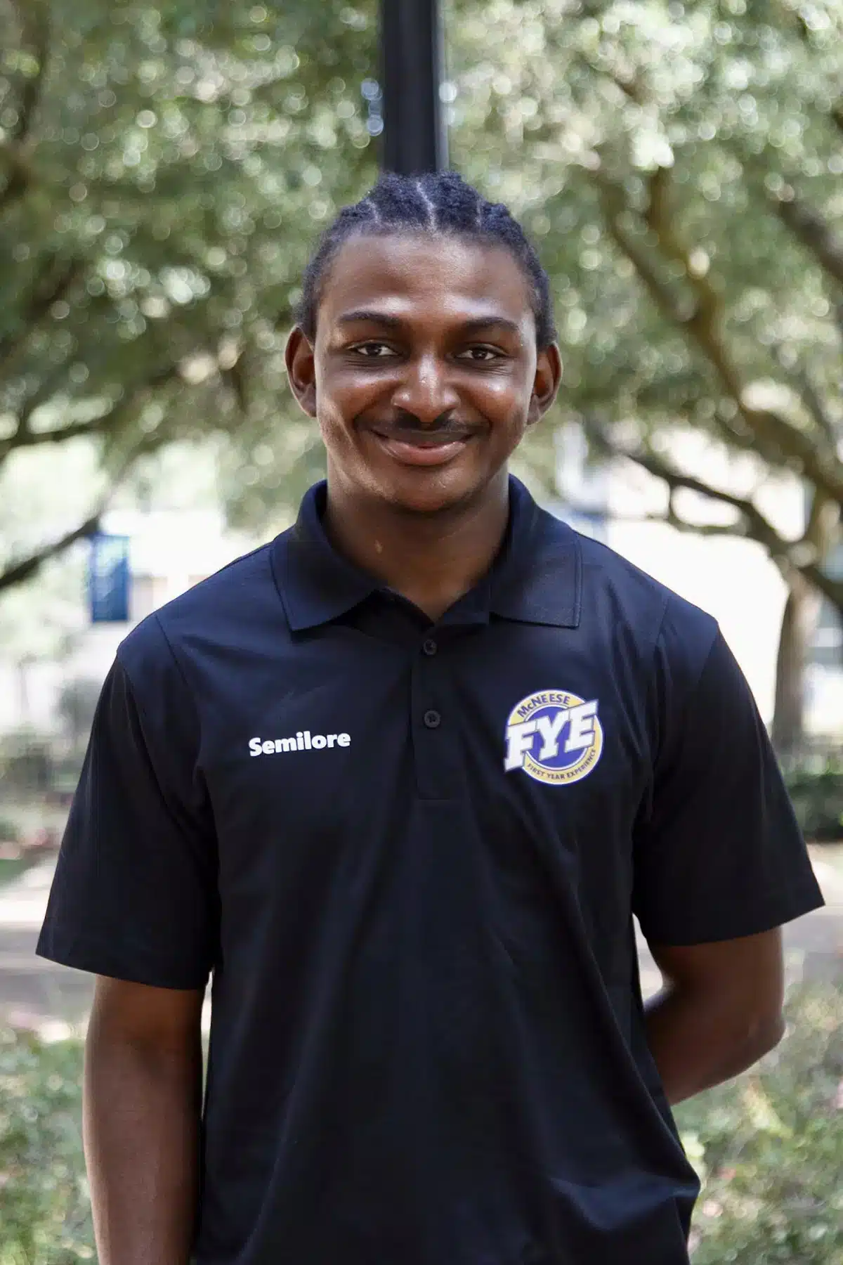 Person smiling outdoors in a black FYE polo shirt with trees in the background.