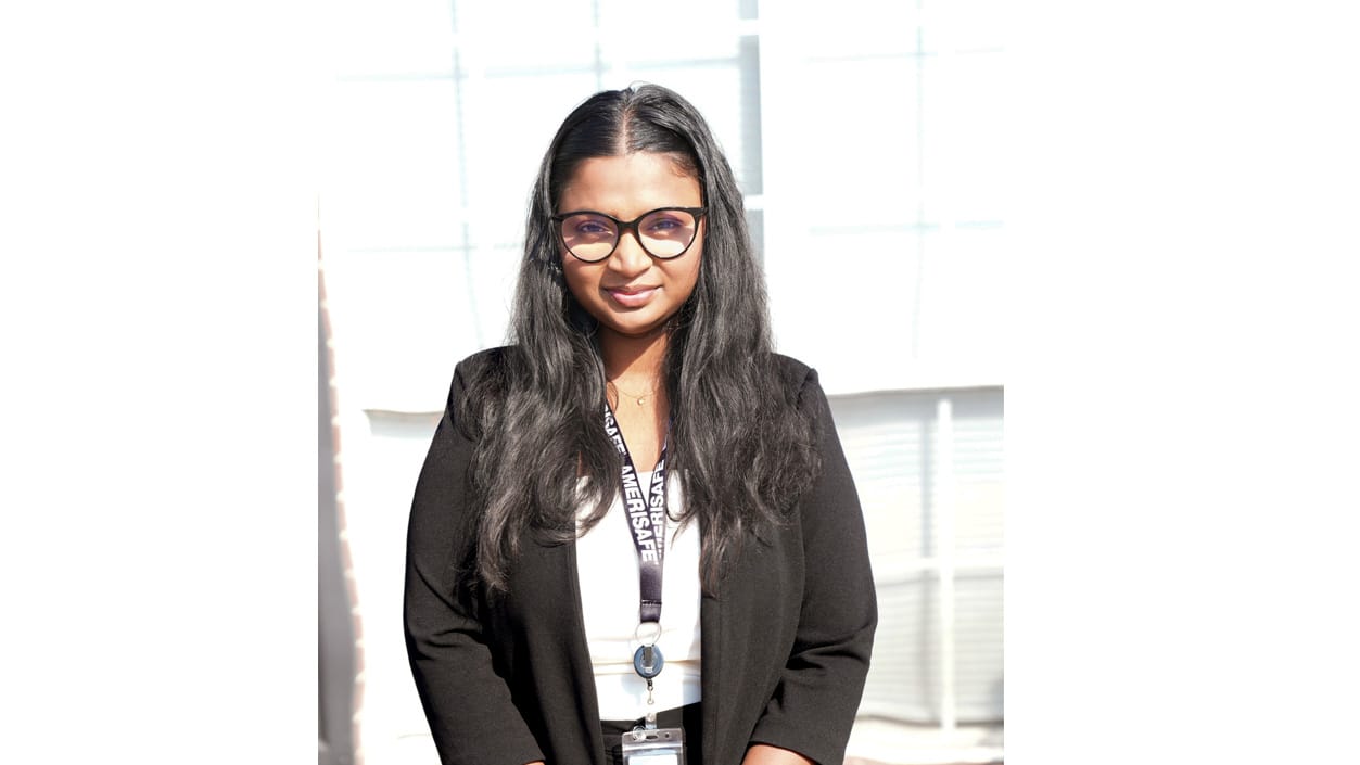 Woman with glasses and long hair wearing a black blazer and lanyard, standing in front of a bright window.