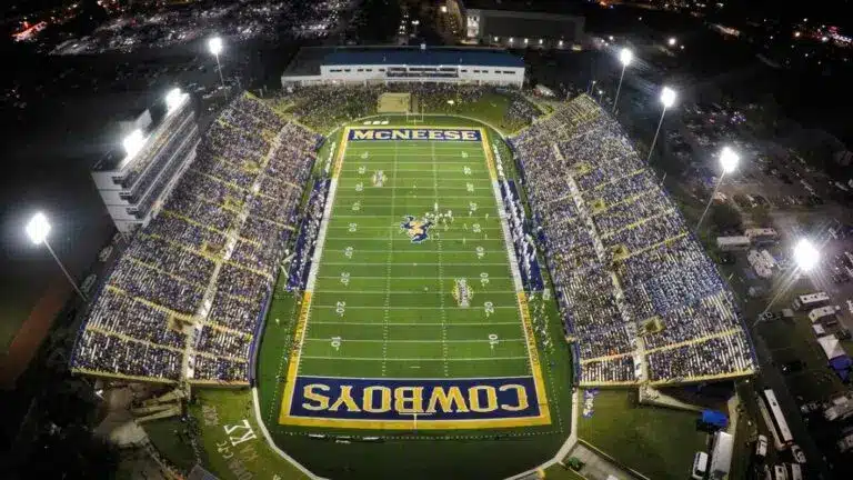 Aerial view of McNeese Cowboys football stadium at night, brightly lit with a full crowd during a game.