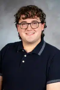 Smiling person with glasses and curly hair wearing a navy polo shirt against a gray backdrop.