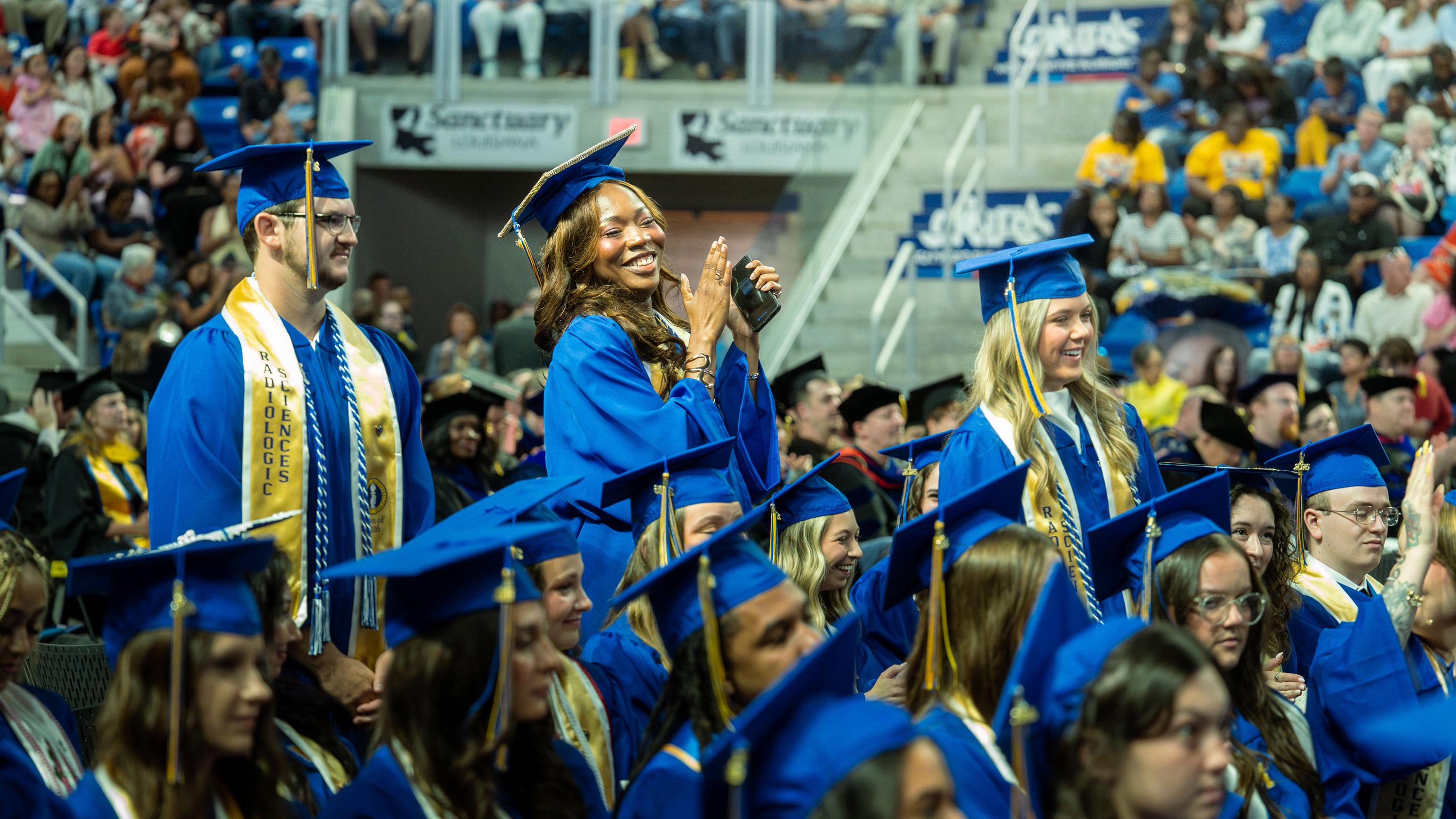 Three people stand to be recognized as honor graduates among McNeese graduates dress in blue graduation regalia.