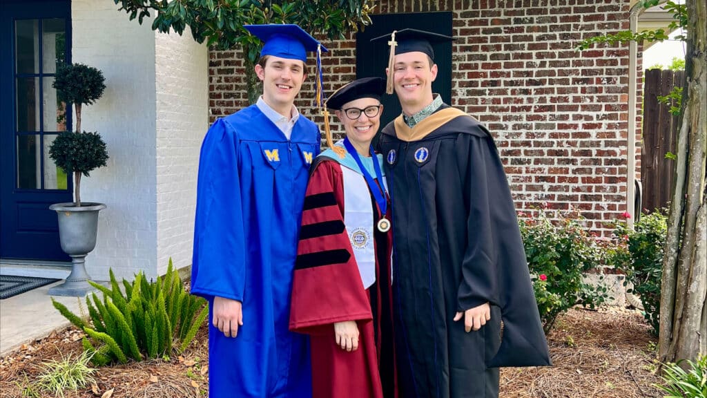 Three people stand together in graduation regalia, a mother and her two sons.