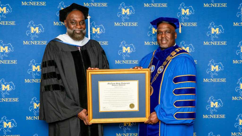 Two men stand in commencement regalia with a framed honorary doctorate degree between them.