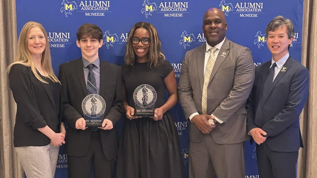 Five people stand in a row smiling at the camera. Two are students who are holding Overall Outstanding Graduate Awards from McNeese.